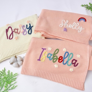 May include: Three personalised baby blankets. One is cream-coloured with the name "Daisy" embroidered in purple, pink, and blue. Two are pink, with the names "Shelly" and "Isabella" embroidered in colourful thread, with flower and rainbow accents.