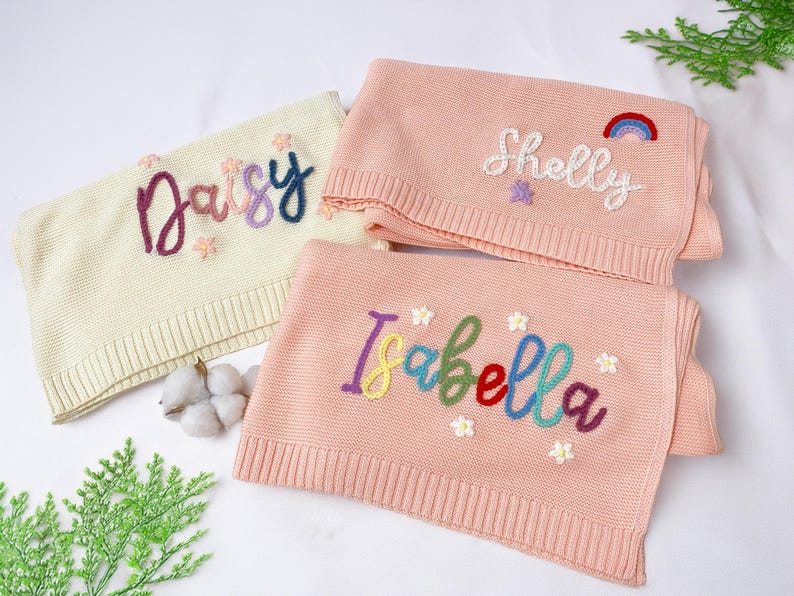 May include: Three personalised baby blankets. One is cream-coloured with the name "Daisy" embroidered in purple, pink, and blue. Two are pink, with the names "Shelly" and "Isabella" embroidered in colourful thread, with flower and rainbow accents.