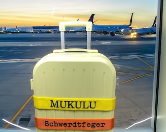 May include: A light yellow rolling suitcase with a retractable handle. The suitcase has two straps with the names "MUKULU" and "Schwerdtfeger" printed on them. The background shows airplanes at an airport.