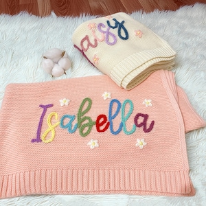 May include: Two knitted baby blankets. One is peach-coloured with the name "Isabella" embroidered in colourful thread, and small flower accents. The other is cream-coloured with the name "Daisy" embroidered in colourful thread, and small flower accents.