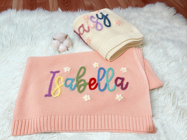 May include: Two knitted baby blankets. One is peach-coloured with the name "Isabella" embroidered in colourful thread, and small flower accents. The other is cream-coloured with the name "Daisy" embroidered in colourful thread, and small flower accents.