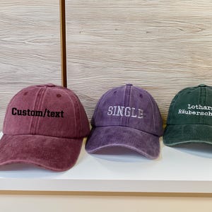 Puede incluir: Tres gorras de béisbol en burdeos, morado y verde. La gorra burdeos tiene bordado "Custom/text". La gorra morada dice "SINGLE". La gorra verde dice "Lothars Räuberscheune".