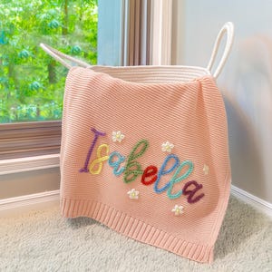 Puede incluir: Una manta de punto rosa claro con el nombre "Isabella" bordado en hilo colorido y pequeños detalles de flores blancas. La manta está sobre una cesta de cuerda blanca con asas, cerca de una ventana.