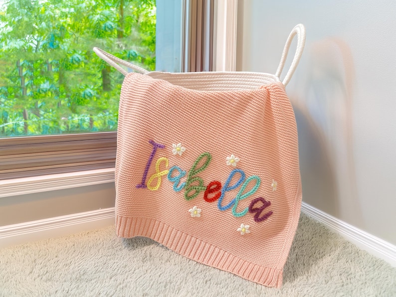 May include: A pale pink knitted blanket embroidered with the name "Isabella" in various colours. The blanket features small white flower details and is draped over a white rope basket. The basket has handles and sits on a light-coloured carpet.