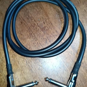 Op de afbeelding: Zwarte instrumentkabel met haakse connectoren. De kabel is opgerold en heeft zilverkleurige stekkers. De kabel is gemarkeerd met "PATENT PENDING LOW NOISE WIRE".
