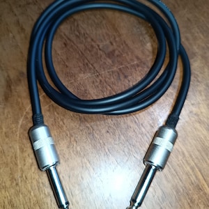 May include: A black audio cable with two silver-colored 1/4 inch (6.35 mm) male connectors. The cable is coiled and lies on a wooden surface. The connectors have a ribbed design.