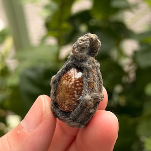 May include: A small, teardrop-shaped pendant featuring a polished, brown and white speckled stone encased in a dark, textured, woven fiber frame. The pendant has a small loop at the top for attachment, showcasing a rustic, organic aesthetic.