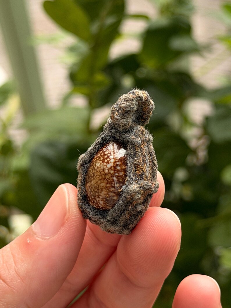 May include: A small, teardrop-shaped pendant featuring a polished, brown and white speckled stone encased in a dark, textured, woven fiber frame. The pendant has a small loop at the top for attachment, showcasing a rustic, organic aesthetic.