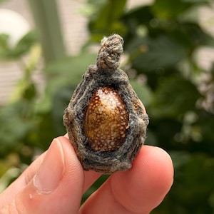 May include: A small, teardrop-shaped pendant featuring a polished, speckled brown and cream-colored stone encased in a textured, dark gray woven fiber frame with a small loop at the top for suspension, showcasing organic patterns and earthy tones.