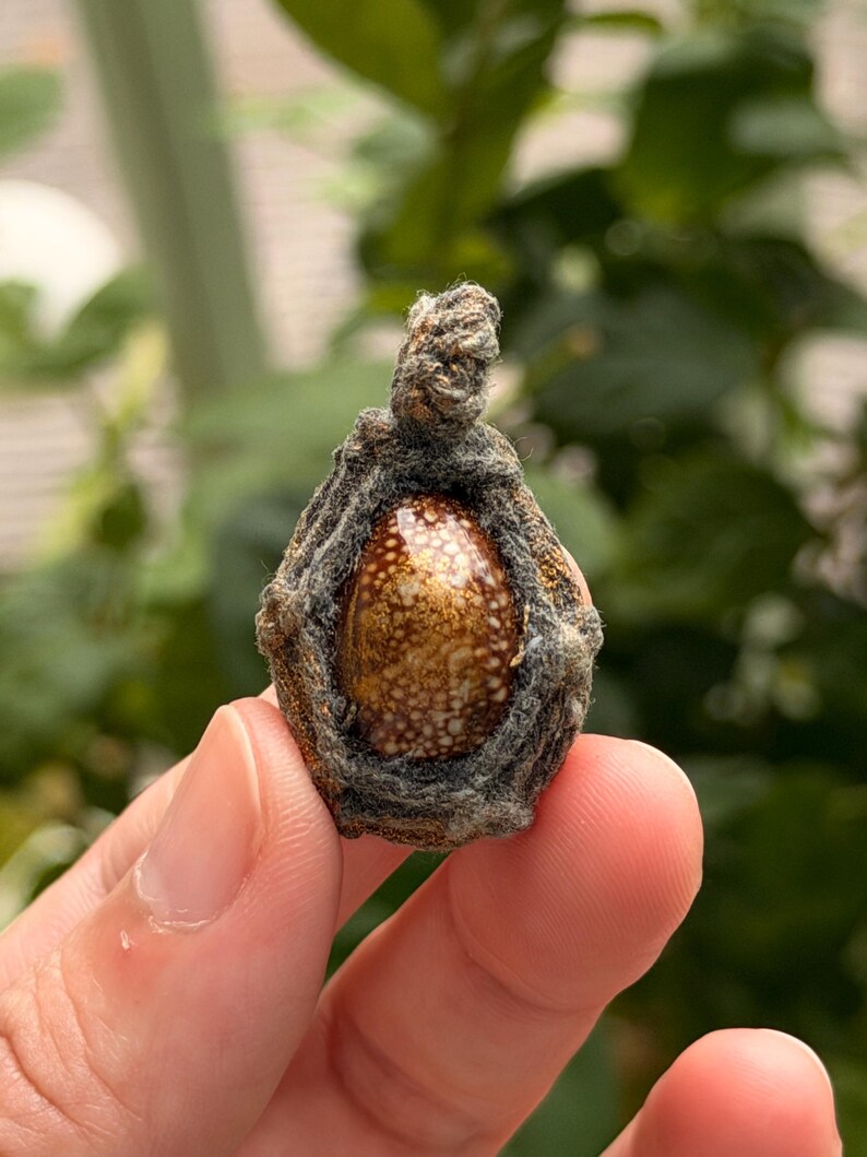 May include: A small, teardrop-shaped pendant featuring a polished, speckled brown and cream-colored stone encased in a textured, dark gray woven fiber frame with a small loop at the top for suspension, showcasing organic patterns and earthy tones.
