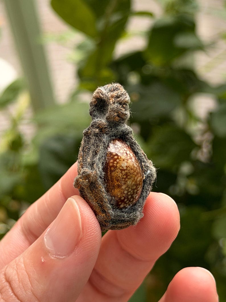 May include: A small, teardrop-shaped pendant featuring a polished, speckled brown and white stone encased in a dark, textured, fibrous woven cord. The pendant has a loop at the top for suspension and a smooth, glossy finish on the central stone.