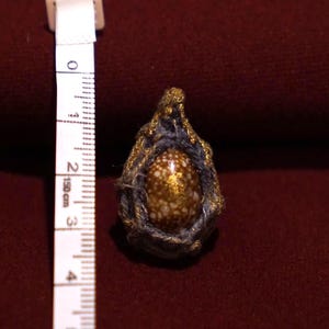 May include: A teardrop-shaped pendant featuring a brown and white speckled shell encased in dark gray fiber with gold-colored accents. A white measuring tape is positioned next to the pendant, showing a height of approximately 1.5 inches.
