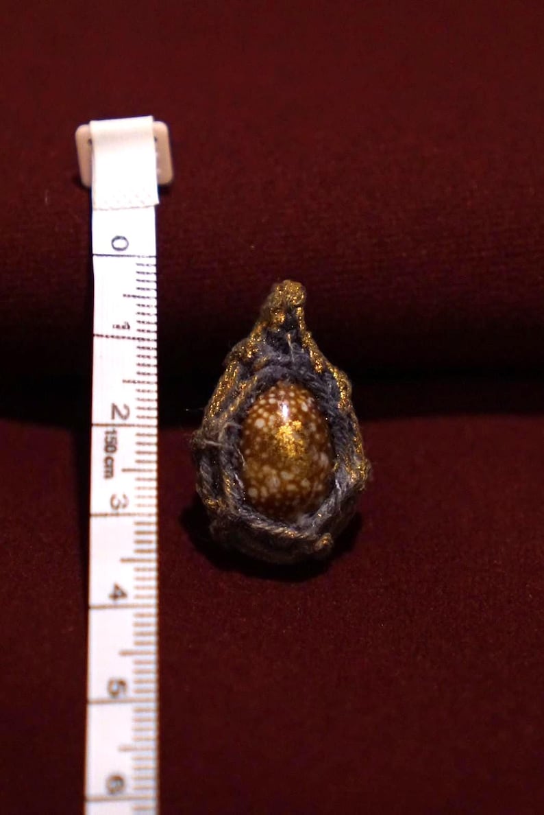May include: A teardrop-shaped pendant featuring a brown and white speckled shell encased in dark gray fiber with gold-colored accents. A white measuring tape is positioned next to the pendant, showing a height of approximately 1.5 inches.
