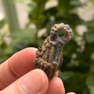May include: A small, dark, textured pendant wrapped in thick, fibrous cord with a circular loop at the top. The surface of the pendant features a weathered, brownish-grey appearance with subtle metallic gold flecks embedded within the woven material.