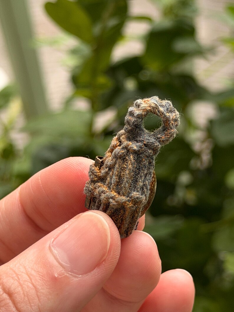 May include: A small, dark, textured pendant wrapped in thick, fibrous cord with a circular loop at the top. The surface of the pendant features a weathered, brownish-grey appearance with subtle metallic gold flecks embedded within the woven material.