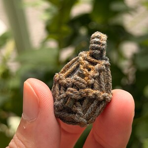 May include: A small, teardrop-shaped amulet encased in a complex, hand-knotted macrame cage made of dark grey and metallic bronze-colored cord. The woven structure features a loop at the top for suspension, with a dense, textured, and knotted pattern.