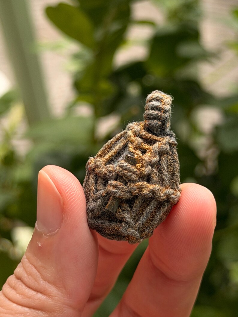 May include: A small, teardrop-shaped amulet encased in a complex, hand-knotted macrame cage made of dark grey and metallic bronze-colored cord. The woven structure features a loop at the top for suspension, with a dense, textured, and knotted pattern.