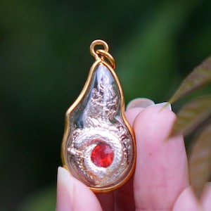 Op de afbeelding: Een goudkleurige hanger met een helder, druppelvormig midden. Binnenin omringt een gedetailleerd zilveren drakenontwerp een grote, ronde, rode edelsteen. De hanger heeft een gouden lus aan de bovenkant voor een ketting.