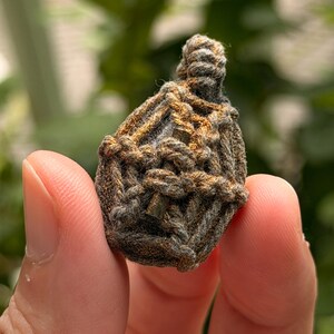 May include: A small, teardrop-shaped amulet encased in a complex, hand-knotted macrame cage made of dark gray and metallic bronze-colored cord. The woven structure features a tight, intricate pattern that secures the central object firmly.