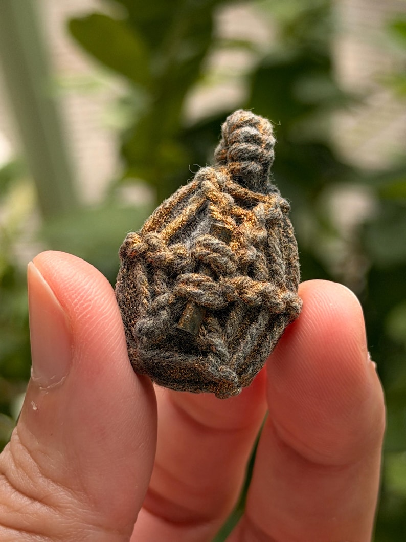 May include: A small, teardrop-shaped amulet encased in a complex, hand-knotted macrame cage made of dark gray and metallic bronze-colored cord. The woven structure features a tight, intricate pattern that secures the central object firmly.