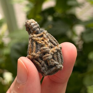 May include: A small, teardrop-shaped amulet encased in a dark gray, textured cord netting with subtle copper-colored highlights. The woven fiber structure features a loop at the top, creating a secure, knotted cage around the central object.