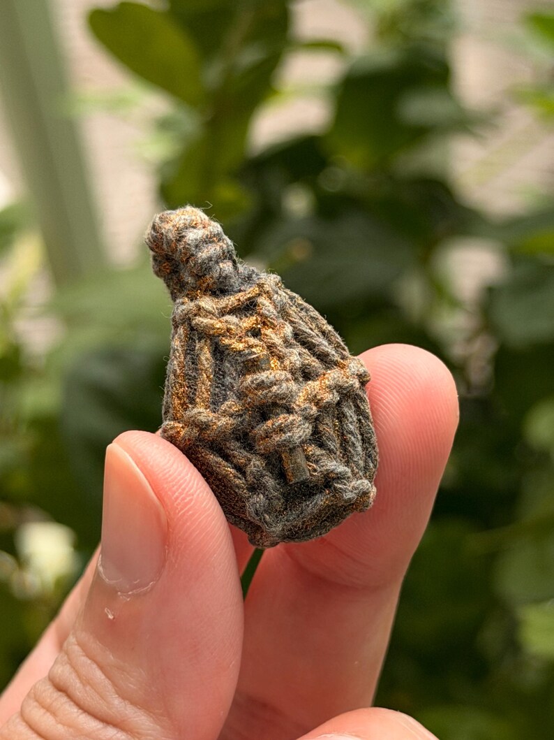 May include: A small, teardrop-shaped amulet encased in a dark gray, textured cord netting with subtle copper-colored highlights. The woven fiber structure features a loop at the top, creating a secure, knotted cage around the central object.