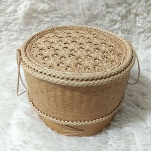 Peut inclure: Un panier rond en osier avec un couvercle. Le panier est fait de fibres naturelles brun clair, avec un bord tressé et un motif décoratif sur le couvercle. Il a deux petites poignées.