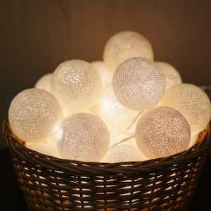 May include: A woven basket filled with glowing, spherical string lights. The lights are a warm white color and are arranged in a cluster. The basket is made of brown wicker and is sitting on a dark surface.