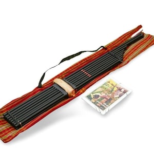 Puede incluir: Un instrumento de panpipe negro con boquilla de madera y correas negras, en un estuche de transporte a cuadros rojos y amarillos. Una pequeña tarjeta rectangular con una imagen colorida está al lado del instrumento.