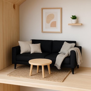 Könnte beinhalten: Eine Miniatur-Wohnzimmerszene mit einem schwarzen Sofa, beigen Kissen und einer grauen Decke. Ein kleiner, runder Holztisch steht auf einem gewebten Teppich. Ein gerahmtes abstraktes Kunstwerk und eine Topfpflanze vervollständigen die Einrichtung.