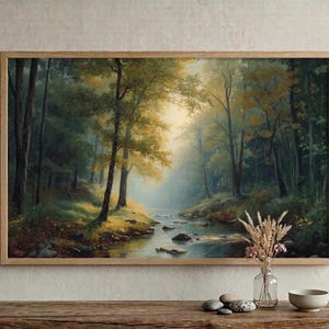 Könnte beinhalten: Gerahmtes Landschaftsgemälde, das eine Waldszene mit einem Fluss zeigt. Das Kunstwerk zeigt grüne Bäume, eine neblige Atmosphäre und goldenes Licht, das durch das Blätterdach scheint. Der Rahmen ist aus hellem Holz. Das Bild strahlt Ruhe aus.