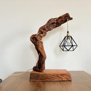 Puede incluir: Una lámpara de mesa de madera única con una base de madera natural y curvada y una pantalla geométrica de metal negro. La lámpara tiene un color marrón cálido y una bombilla decorativa. La lámpara está sobre una mesa de madera.