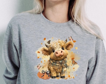 May include: A grey sweatshirt with a charming illustration of a Highland cow calf. The calf is wearing a scarf and is surrounded by pumpkins, butterflies, and autumn leaves. The design has a watercolour effect.