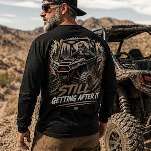 May include: Black long-sleeve shirt with a graphic of an off-road vehicle and the text "STILL GETTING AFTER IT." The shirt features a crew neck and is made of a soft material. The image is set against a desert backdrop.