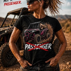 May include: Black t-shirt with a graphic of a pink and black off-road vehicle kicking up dirt. The text "NOT A PASSENGER" and "PASSENGER" with a red X are printed on the shirt. The image is set in a desert landscape.