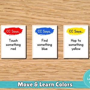 May include: Three rectangular cards with color-based instructions: "Touch something red," "Find something blue," and "Hop to something yellow." The cards are on a wooden surface, with the text "Move & Learn Colors" below.