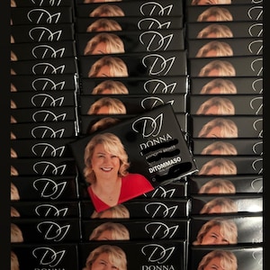 May include: A collection of black boxes, each displaying a smiling woman in a red top. The boxes are labelled with the name "DONNA" and "DITOMMASO". The boxes are stacked in a neat arrangement.