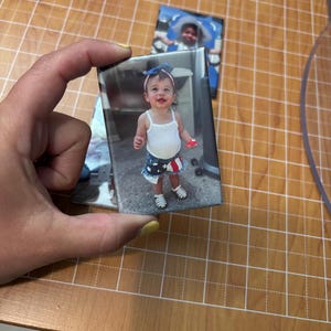 May include: A rectangular photo print featuring a smiling baby girl in a white tank top and a patriotic skirt with red, white, and blue stars. The baby is wearing a blue bow and white shoes. Another photo is visible in the background.
