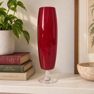 May include: A tall, cylindrical, deep red glass vase with a smooth, glossy finish. The vase features a clear, twisted glass stem that connects to a circular, transparent glass base, providing a stable foundation for the elongated vessel.