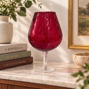 May include: A deep red glass vase with a textured, dimpled surface and a wide, rounded body. The vase features a clear, twisted glass stem and a circular base, standing upright with a scalloped rim at the top of the vessel.