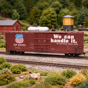 Puede incluir: Maqueta de un vagón de mercancías Union Pacific de color granate, con el logotipo y el texto "We can handle it." El modelo está sobre vías, con árboles, edificios y una torre de agua de fondo.