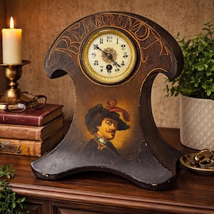 May include: A dark brown wooden mantel clock with a gold-colored clock face. The clock features a portrait of a man in a hat and the name "Rembrandt" above the clock face. The clock is on a wooden surface.