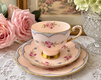 May include: A vintage tea set featuring a teacup, saucer, and dessert plate. The set is primarily pink with a light blue trim and gold accents. Floral patterns of pink roses and blue accents decorate the set, creating a delicate and elegant aesthetic.