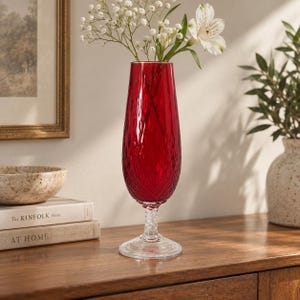 Puede incluir: Un jarrón de cristal rojo vibrante y alto con una superficie texturizada de patrón de diamantes. El jarrón cuenta con un tallo de cristal transparente retorcido y una base circular de cristal transparente. Contiene flores blancas y un lirio.