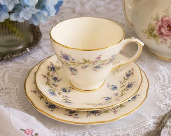 May include: Cream-colored teacup, saucer, and side plate set with gold trim and delicate blue floral patterns. A matching teapot with a pink rose design is visible in the background. The set is arranged on a white lace tablecloth.