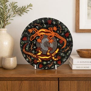Puede incluir: Un plato decorativo con fondo negro que presenta dos lagartos naranjas y amarillos que rodean un diseño central. El plato tiene un diseño artístico único con un patrón de puntos y se exhibe sobre un soporte blanco.