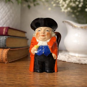 May include: A vintage ceramic figurine of a man in a black hat and orange coat, holding a small yellow glass. The figurine has a blue vest, white collar, and black pants. It is a decorative item, likely for display.
