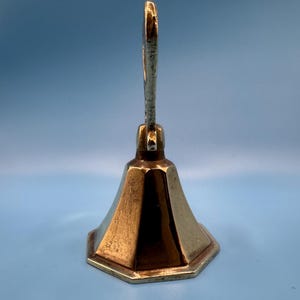 K&ouml;nnte beinhalten: Eine Vintage-Messingglocke mit einem langen, gebogenen Griff und einem ausgestellten, sechseckigen Sockel. Die Glocke hat einen warmen, goldenen Farbton und ein schlichtes, elegantes Design. Der Griff ist oben an der Glocke befestigt.