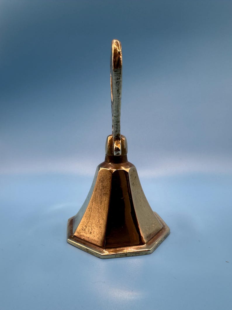 K&ouml;nnte beinhalten: Eine Vintage-Messingglocke mit einem langen, gebogenen Griff und einem ausgestellten, sechseckigen Sockel. Die Glocke hat einen warmen, goldenen Farbton und ein schlichtes, elegantes Design. Der Griff ist oben an der Glocke befestigt.