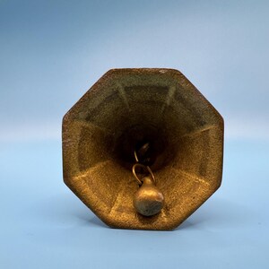 K&ouml;nnte beinhalten: Vintage Messingglocke mit achteckiger Oberseite und kleinem Kl&ouml;ppel. Die Glocke hat ein angelaufenes, gealtertes Aussehen mit einem gr&uuml;nlichen Farbton in einigen Bereichen. Der Hintergrund ist einfarbig hellblau.